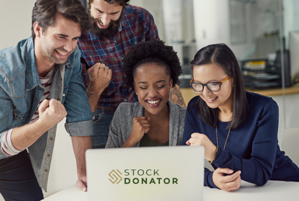 Group of diverse individuals celebrating around a laptop displaying the Stock Donator logo, emphasizing teamwork and engagement in stock donation initiatives for nonprofits.